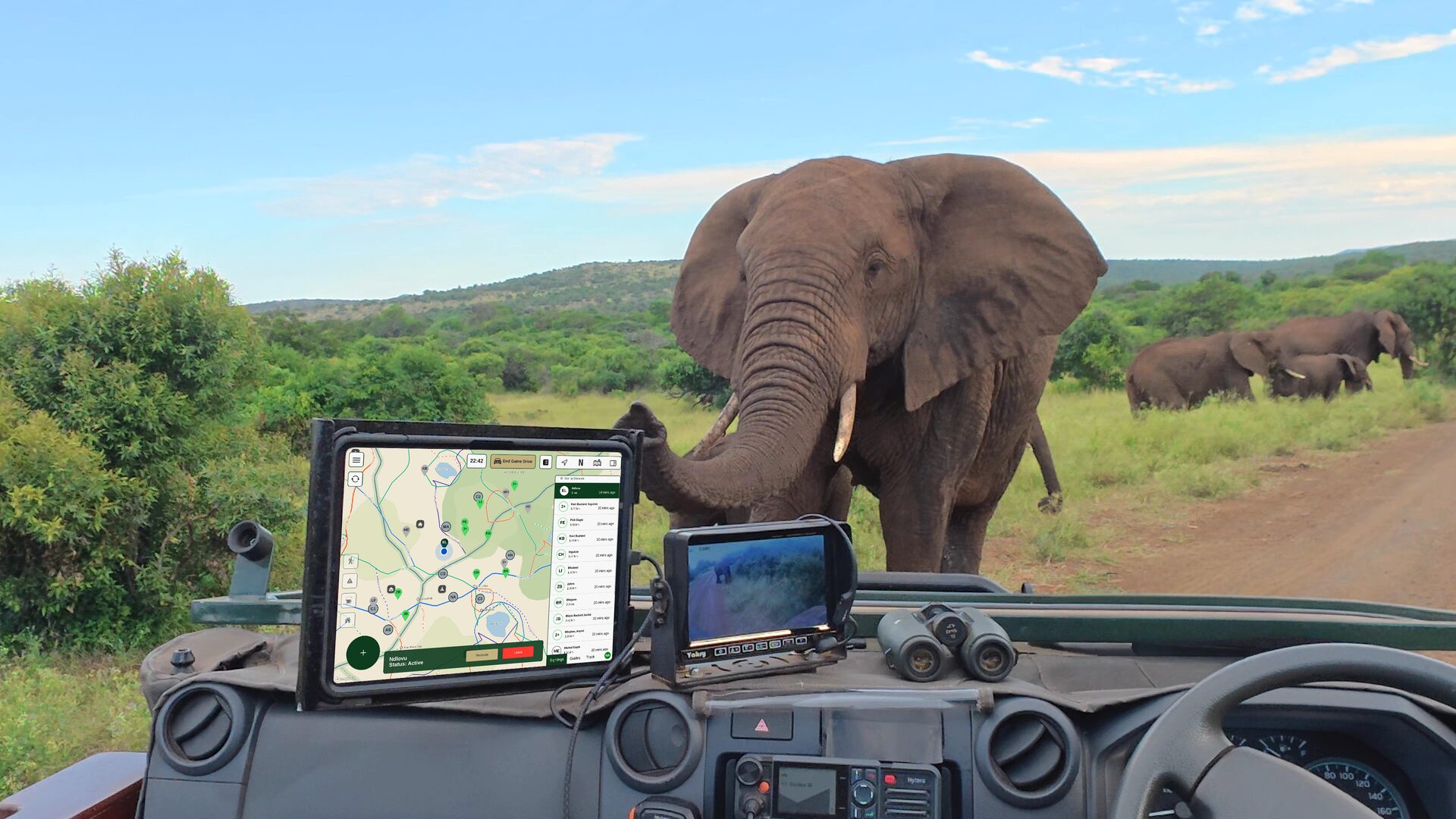A tablet mounted on the dashboard of a game viewer shows the WildSight live reserve map with sighting pins, while a bull elephant crosses the road ahead and a breeding herd feeds in the bush beyond.